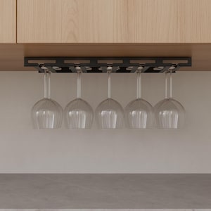 May include: A black metal wine glass rack installed under a wooden cabinet, holding five clear wine glasses. The glasses are suspended upside down, showcasing their rounded bowls and slender stems. The rack is designed to hold multiple glasses.