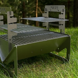 May include: A silver metal barbecue with the word "NAME" on the side. The barbecue has a rectangular firebox, a cooking grate, and a warming rack. It is set up on grass.