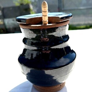 May include: A handmade ceramic jar with a dark blue glaze and a wooden handle. The jar has a lid and a unique shape, with a brown base and rim. The jar is likely used for storing food or other items.