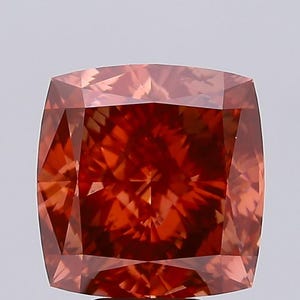 May include: A cushion-cut diamond with a warm, reddish-orange hue. The gemstone's facets reflect light, creating a brilliant display. The square shape with rounded corners is a classic design for engagement rings and fine jewelry.