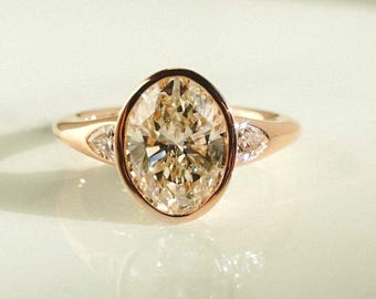 May include: Rose gold engagement ring featuring a large, oval-cut diamond center stone. The diamond is surrounded by a bezel setting and flanked by two smaller, triangular diamonds. The ring has a polished finish and a classic design.