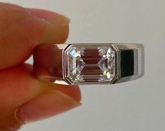 May include: A silver-toned metal ring featuring a large, rectangular emerald-cut clear gemstone set horizontally in the center. The band is wide and smooth with a polished finish, showcasing a clean, geometric design that highlights the stone.