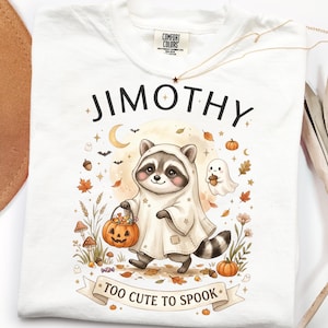 以下が含まれることがあります： 白いTシャツに、お化けの仮装をしてジャック・オー・ランタンのバケツを持ったアライグマのグラフィックが描かれています。JIMOTHYとTOO CUTE TO SPOOKという文字があり、秋の葉、キノコ、星、小さな幽霊のイラストが周囲に配置されています。