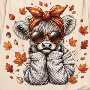 May include: A beige t-shirt featuring a graphic of a fluffy Highland cow wearing a patterned headscarf, oversized sunglasses, and a textured sweater, surrounded by scattered autumn leaves and acorns in shades of orange, brown, and yellow.