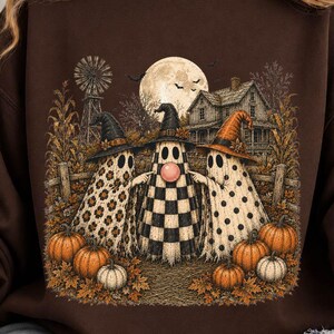 May include: A dark brown crewneck sweatshirt featuring a graphic print of three ghosts wearing witch hats standing in a field with pumpkins. The ghosts have patterns of leopard print, black and white checkers, and polka dots, with a full moon background.