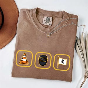 May include: Brown Comfort Colors t-shirt with three yellow-bordered icons: a traffic cone, a welding mask, and a skull and crossbones flag. A silver necklace and a brown hat are also visible.