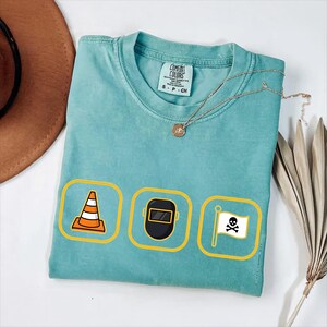 May include: A turquoise Comfort Colors t-shirt featuring three yellow-bordered icons: an orange traffic cone, a black welding mask, and a white pirate flag with a skull and crossbones. A gold necklace and a brown hat are also visible.