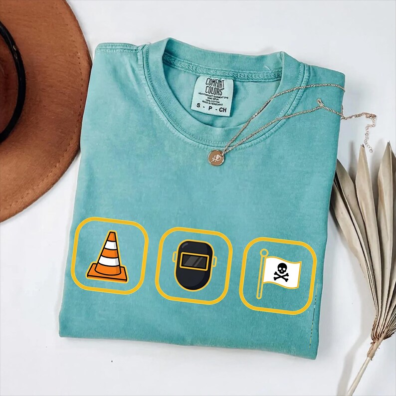 May include: A turquoise Comfort Colors t-shirt featuring three yellow-bordered icons: an orange traffic cone, a black welding mask, and a white pirate flag with a skull and crossbones. A gold necklace and a brown hat are also visible.