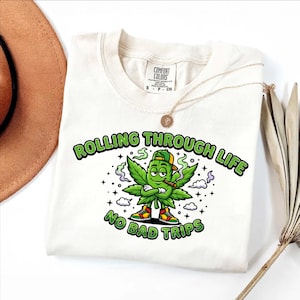 May include: White t-shirt with a graphic of a cannabis leaf character wearing a green cap, smoking a cigar, and wearing sneakers. The text "Rolling Through Life, No Bad Trips" is printed above and below the character in green.