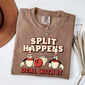 Può includere: T-shirt Comfort Colors marrone con la scritta "SPLIT HAPPENS DEAL WITH IT" in rosso e bianco. La grafica presenta birilli da bowling e una palla da bowling. Sono visibili anche una collana d'argento e un cappello marrone.