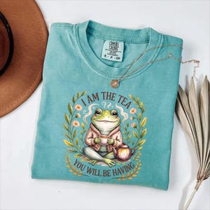 May include: A turquoise Comfort Colors t-shirt featuring a frog illustration. The frog is wearing a pink jacket and holding a teacup, with the text "I AM THE TEA YOU WILL BE HAVING". A gold necklace and a brown hat are also visible.