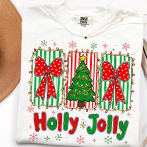 May include: A white t-shirt featuring a graphic print of three rectangular panels with green and red stripes. The outer panels display large red bows with white polka dots, while the center panel shows a green Christmas tree with a yellow star.
