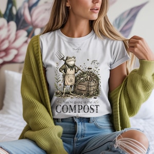 Op de afbeelding: Wit t-shirt met een kikkerillustratie met een hooivork en naast een compostbak. De tekst op het shirt luidt "That's it. You're going in the COMPOST."