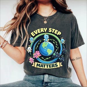 May include: Dark gray t-shirt featuring a colorful graphic design. The design includes the text "EVERY STEP MATTERS" with a depiction of the Earth surrounded by cartoon characters. The shirt is a casual wear item.