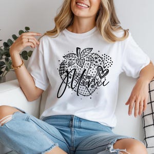 Può includere: T-shirt bianca con grafica in bianco e nero di una zucca e la scritta "Mami". Il design della zucca incorpora vari motivi, tra cui pois e quadri. Nel design è inclusa una forma di cuore. Abbigliamento casual.