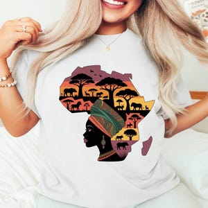 May include: White t-shirt featuring a colorful graphic of the African continent filled with silhouettes of trees, animals, and a woman's profile. The design incorporates shades of orange, purple, and green, creating a vibrant and artistic representation of Africa.