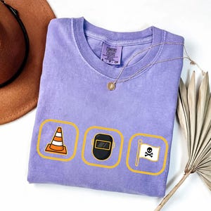 May include: A lilac Comfort Colors t-shirt with three yellow-bordered icons: a traffic cone, a welding mask, and a skull and crossbones flag. A gold necklace and a brown hat are also visible.