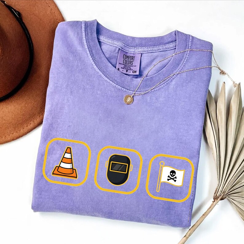 May include: A lilac Comfort Colors t-shirt with three yellow-bordered icons: a traffic cone, a welding mask, and a skull and crossbones flag. A gold necklace and a brown hat are also visible.