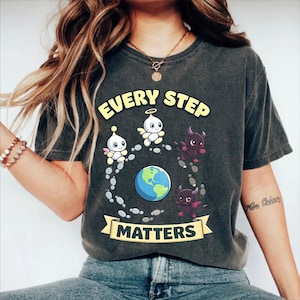 May include: Dark gray t-shirt with the text "EVERY STEP MATTERS" in yellow. The shirt features a cartoon design of angel and devil characters surrounding a globe. The design is printed on a soft, comfortable fabric.