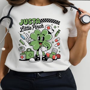 Puede incluir: Camiseta blanca con un personaje de trébol de dibujos animados con gorro de enfermera, rodeado de suministros médicos. El texto "Just a Little Pinch" está encima del trébol. El trébol es verde, con zapatos blancos y negros. Incluye jeringas, pastillas y un botiquín.
