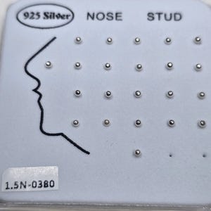 May include: A display of silver nose studs on a white card. The card has the text "925 Silver", "NOSE STUD", and a profile outline. The studs are small, round, and evenly spaced. The text "1.5N-0380" is also visible.