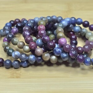 May include: Several beaded bracelets in shades of purple, blue, and tan are arranged on a wooden surface. The bracelets are made of round beads, each approximately 0.3 inches in diameter, and are strung together to form a circular shape.