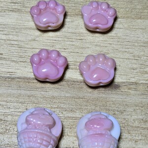 May include: A collection of six pink resin beads. Four beads are paw prints, and two are bunny figures in baskets. The beads are light pink with a glossy finish. The background is a light brown wooden surface.
