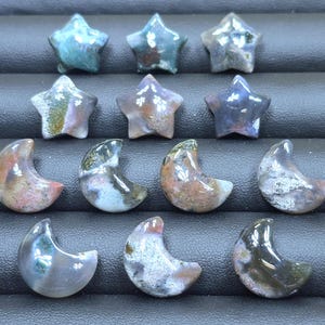 May include: A collection of star and crescent moon shaped gemstones in various colors, including green, brown, and purple. The gemstones are arranged on a black surface, showcasing their unique patterns and textures.