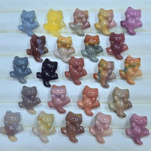 May include: A collection of twenty-five small, carved stone cat figurines arranged in five rows. Each cat features a smooth, polished finish and displays a unique color, including shades of blue, yellow, brown, black, pink, and multi-colored patterns.