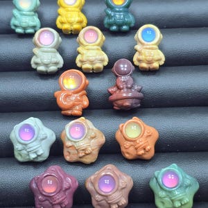 May include: A collection of small, colorful astronaut figurines and star-shaped objects. The astronaut figures are in various colors, including green, yellow, and orange, with glowing, round visors. The star-shaped objects have a similar glowing center.