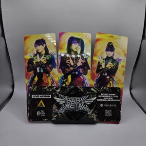 May include: Three BABYMETAL concert tickets in a black stand. Each ticket features a different member in colourful outfits. The bottom ticket has the BABYMETAL logo, Live Nation logo, and concert details: Intuit Dome, November 1, 2025.