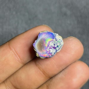 May include: A raw, unpolished opal gemstone displaying a vibrant play-of-color with flashes of neon blue, green, and red against a translucent, milky white base. The stone has an irregular, organic shape with a rough, textured outer edge.