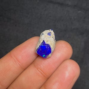 May include: A raw, natural opal gemstone with a vibrant blue color. The opal is encased in a rough, beige matrix, showcasing its natural form. The gemstone has a polished, glossy appearance, with hints of green and blue.