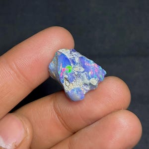 May include: A rough, uncut opal gemstone with a vibrant display of colors. The opal features flashes of blue, pink, and green within a matrix of white and gray. The gemstone is held between two fingers against a dark background.