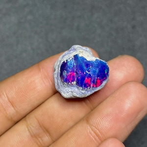 May include: A raw, unpolished opal gemstone featuring a rough, light grey outer crust and a vibrant, polished-looking centre displaying intense flashes of electric blue, deep purple, and bright neon pink colours across its irregular surface.