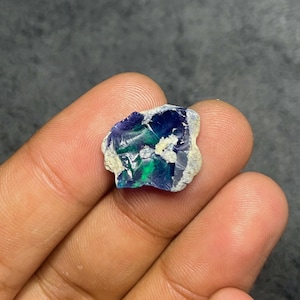 May include: A raw opal gemstone displaying a play of colour. The stone exhibits flashes of blue, green, and purple within a light grey matrix. The opal is held in a hand, highlighting its natural, unpolished state.