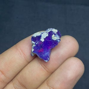 May include: A raw gemstone with a striking deep purple colour, interspersed with white patches and touches of red. The stone's irregular shape and natural texture are visible, held in a hand.