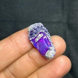 Könnte beinhalten: Ein roher Opal-Edelstein in Lila- und Blautönen. Der Stein hat eine raue, strukturierte Oberfläche mit weißen Bereichen und einem rosa Schimmer. Dieser natürliche Edelstein eignet sich für die Schmuckherstellung oder zum Sammeln. Etwa 3 cm lang.