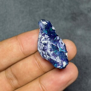 May include: A raw, unpolished gemstone featuring a deep indigo and violet base colour with iridescent flashes of bright green and blue throughout its textured, jagged surface. The stone displays a natural, irregular shape with crystalline inclusions.