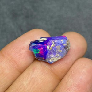 Könnte beinhalten: Ein roher, unpolierter Opal-Edelstein mit einem lebendigen Farbspiel aus tiefem Blau, Violett und leuchtendem Neonpink. Der Stein hat eine unregelmäßige, gezackte Form mit einer strukturierten, felsigen Oberfläche und einem durchscheinenden Aussehen.