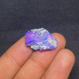Puede incluir: Una vibrante piedra preciosa de ópalo en bruto con una mezcla de colores, incluyendo púrpura, azul, rosa y toques de verde. La piedra en bruto tiene una superficie texturizada y se sostiene en una mano. El ópalo es una gema natural sin cortar.