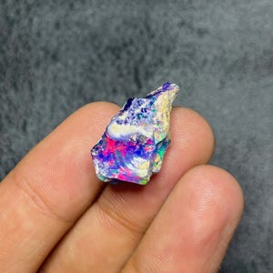 May include: A colourful, raw opal gemstone displaying pink, blue, green, and white hues. The rough stone is held in a hand, highlighting its natural, unpolished texture and iridescent play of colour. The opal's surface reflects light, creating a dazzling display.