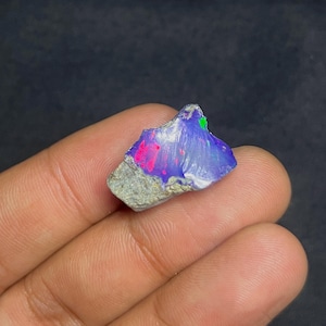 May include: A vibrant, raw opal gemstone with a mix of colors. The stone displays a striking play of color, with flashes of pink, blue, and green. The opal is held in a hand, showcasing its natural, rough texture and unique beauty.