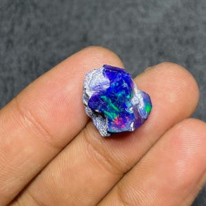 May include: A raw, unpolished opal gemstone featuring a dark, textured matrix base with vibrant flashes of iridescent blue, green, and purple color play across its surface, showcasing a natural, irregular shape and a multifaceted, reflective appearance.