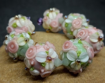 Könnte beinhalten: Eine Reihe von Glasperlen mit Blumenmuster. Die Perlen sind in den Farben Rosa, Weiß und Grün.