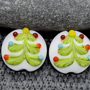 May include: Two round, white glass Christmas tree ornaments. Each ornament features a green Christmas tree with colorful, round ornaments. The trees have yellow toppers, red, orange, and blue ornaments. The ornaments are on a dark gray surface.