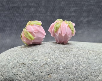 Könnte beinhalten: Zwei handgefertigte, rosa Glasblumenskulpturen mit grünen Akzenten. Die zarten Blütenblätter sind zartrosa und die Blätter leuchtend grün. Die Blumen werden auf einer grauen Steinoberfläche ausgestellt.