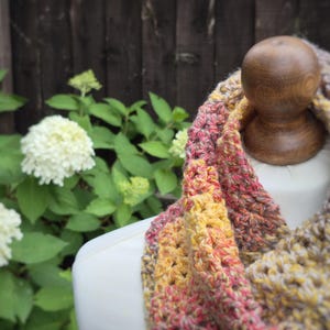 May include: A handmade crochet infinity scarf in shades of yellow, pink, and brown, displayed on a mannequin. The scarf has a textured appearance and is set against a backdrop of green foliage and a wooden fence.