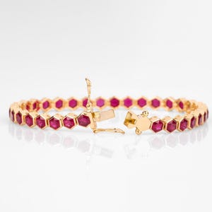 Könnte beinhalten: Ein goldenes Armband mit einer Reihe von sechseckigen, tiefroten Edelsteinen. Das Armband ist geöffnet und zeigt den Verschlussmechanismus. Die Edelsteine sind in einem goldenen Metallband gefasst.