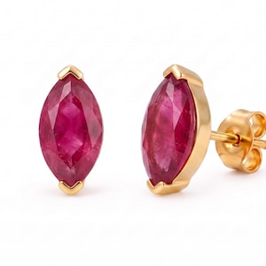 Puede incluir: Un par de pendientes de botón con rubíes rojos de talla marquesa engastados en metal dorado. Un pendiente se muestra de frente, mientras que el otro está inclinado para mostrar el poste dorado y el cierre de mariposa. Las piedras son de color carmesí.